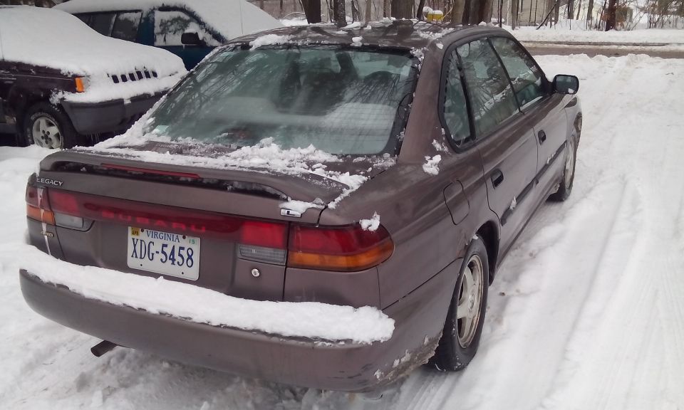 Brown 1996 Subaru Legacy sedan parked in snow, rear view. Another vehicle covered in snow is to the left
