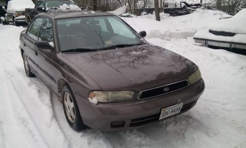 1996 Subaru Legacy - A 1996 Subaru Legacy sedan sits covered in snow, with other vehicles partially visible in the background.