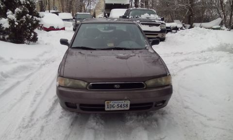 1996 Subaru Legacy - A brown sedan sits in a snow-covered driveway, with other vehicles visible in the background.