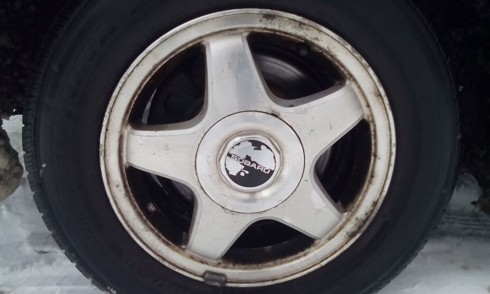 Silver Subaru wheel with a black tire, covered in dirt and grime, with a Subaru logo in the center
