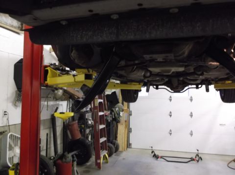 1999 Mercury Grand Marquis - The undercarriage of a vehicle is shown elevated on a lift in a garage setting with various tools and equipment around.