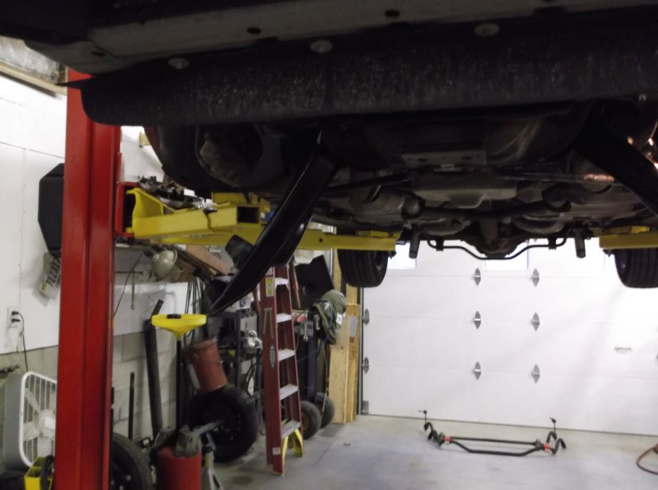 Underside of a car on a lift, showing suspension and exhaust components. Tools and a ladder are visible in the background
