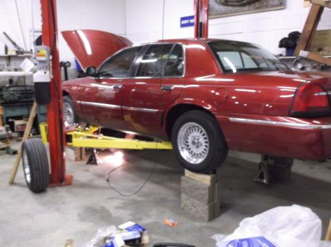 1999 Mercury Grand Marquis - A red 1999 Mercury Grand Marquis is raised on a car lift in a garage, with its hood open and a tire removed.