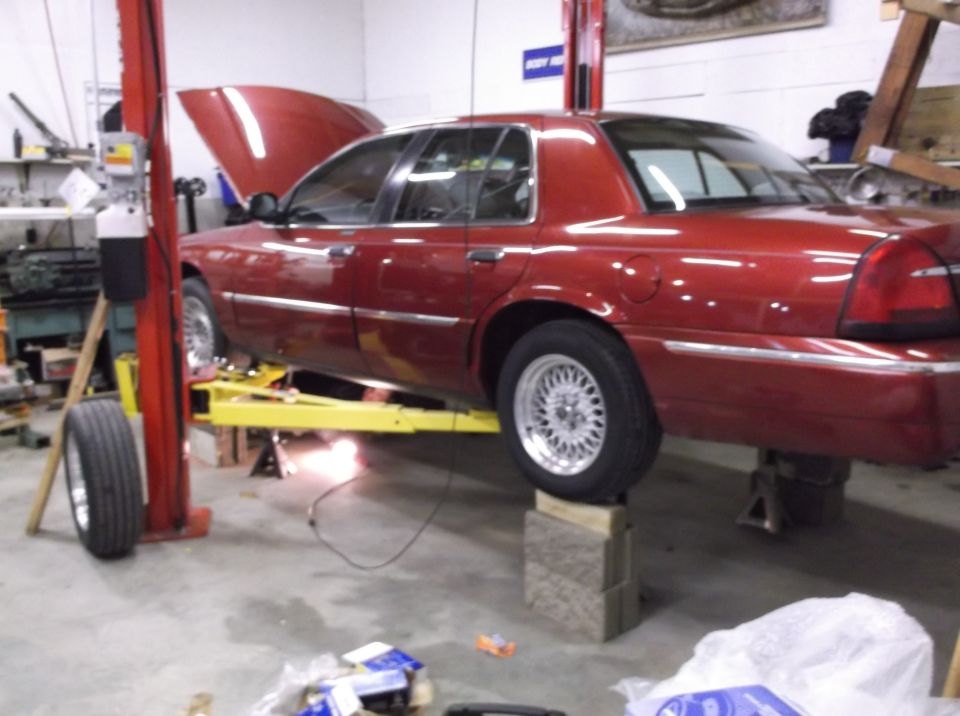 Red 1999 Mercury Grand Marquis on a yellow car lift, hood open, with a detached front tire on the left