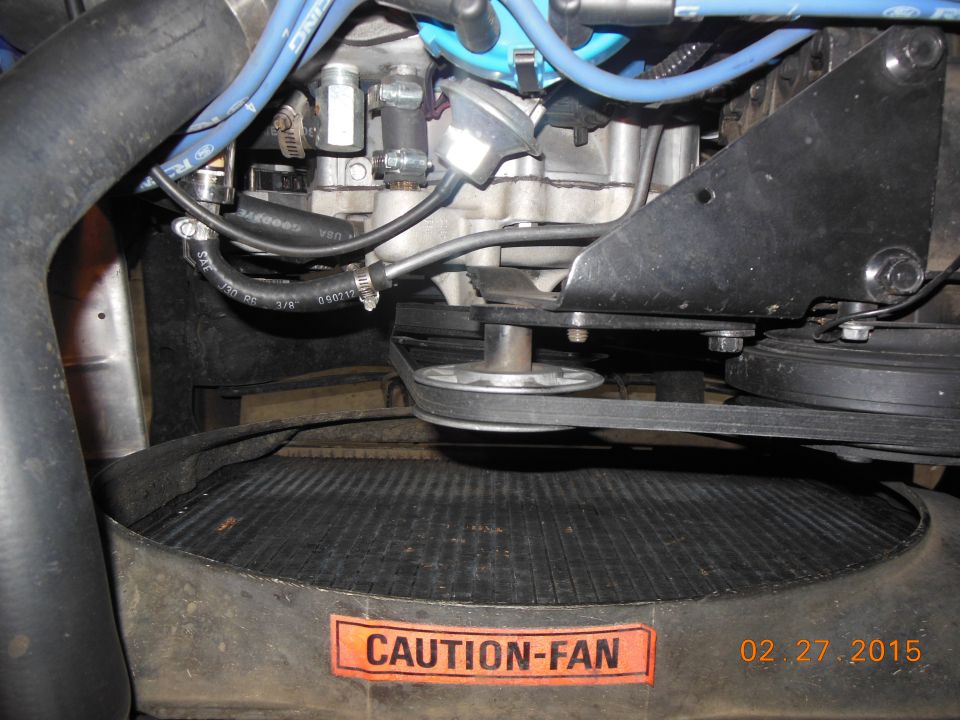 Close-up of engine bay showing fan, belts, and pulleys. Orange sticker says CAUTION-FAN
