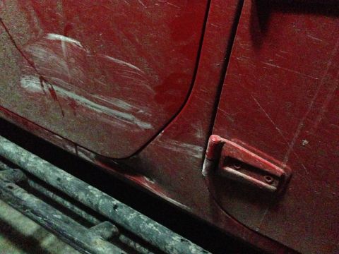 1979 Ford Bronco - Close-up of a red vehicle door showing extensive scratches and damage, with a metal side step visible below.