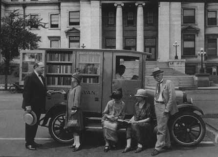 1909 Ford Model T - People gather around a mobile library van parked in front of a large building.