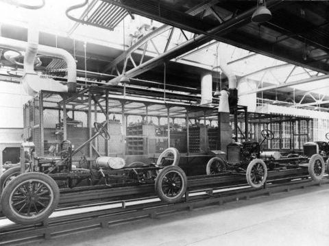 1909 Ford Model T - Early automotive assembly line shows multiple chassis moving along a production line.