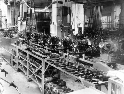 1909 Ford Model T - A factory assembly line is filled with engines and parts, suggesting mass production of vehicles.