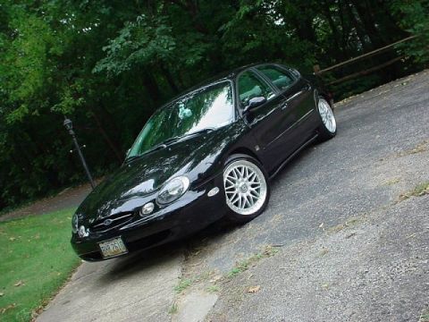 1999 Ford Taurus - A black hatchback is parked on a sloped driveway surrounded by greenery.