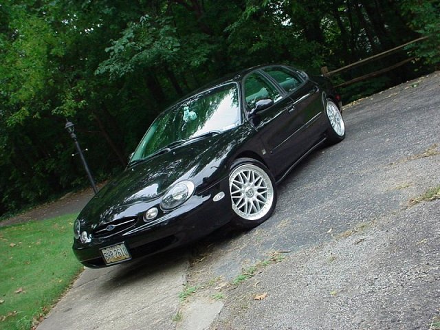 Black Ford Taurus sedan front three-quarter view on a wooded driveway with silver alloy wheels