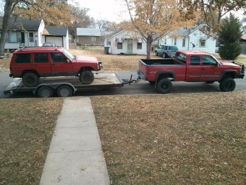 1986 Ford Bronco - A red pickup truck tows a red Jeep Cherokee on a trailer down a residential street.
