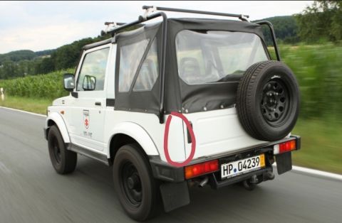1987 Suzuki Samurai - This Suzuki Samurai is seen from the rear, driving on a paved road with fields and trees in the background.