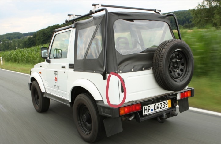 White Suzuki Samurai driving on a road. Has a spare tire on the back and a roof rack