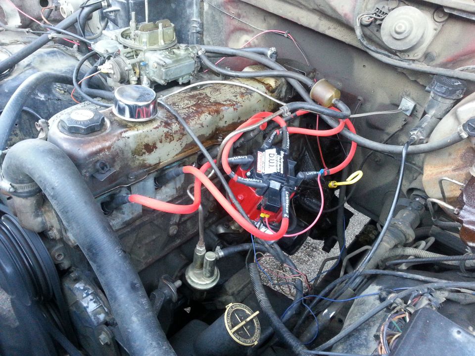Engine bay with carburetor, distributor, and various hoses and wires. Some components look rusty