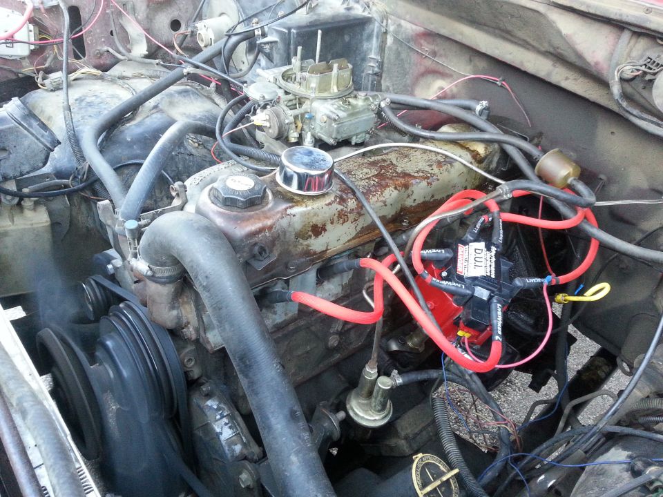 Engine bay with a V8 engine, carburetor, distributor, and hoses. Some rust is visible on the engine block