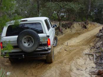 1992 Ford Bronco - A white 1992 Ford Bronco navigates a sandy, off-road trail surrounded by trees.