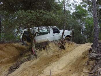 1992 Ford Bronco - A white SUV navigates a steep dirt incline surrounded by trees. The Supermotors.net logo is visible.