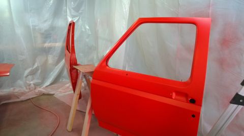 1983 Ford Bronco - A red car door and fender are being painted in a workshop. The parts are mounted on sawhorses.