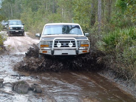 1992 Ford Bronco - A white 1992 Ford Bronco navigates a muddy trail, kicking up water and dirt.