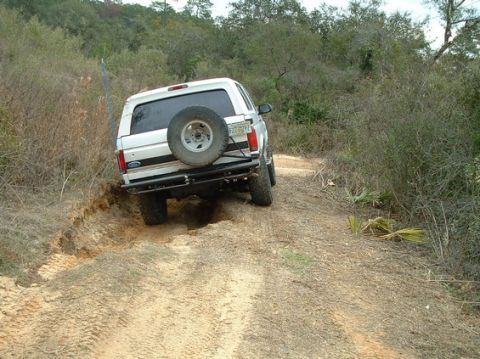 1992 Ford Bronco - A white 1992 Ford Bronco navigates a challenging off-road trail, kicking up dirt as it goes.
