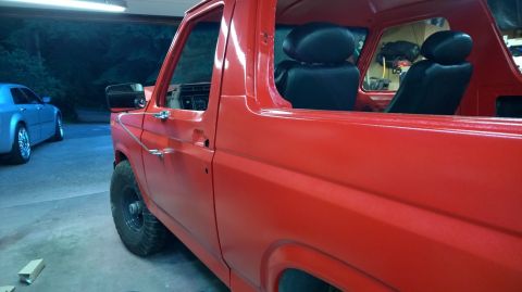 1983 Ford Bronco - Side view of a red 1983 Ford Bronco, revealing its interior with black seats and dashboard, parked next to another vehicle.