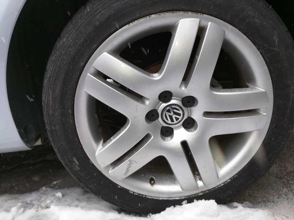 Silver five-spoke Volkswagen wheel with a black tire, showing a VW logo in the center. The car is light-colored