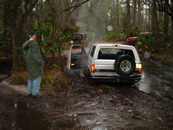 White 1992 Ford Bronco stuck in mud on wooded trail, door open. Another Jeep and red SUV behind