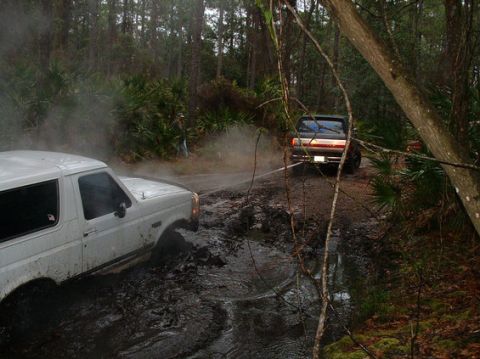 1992 Ford Bronco - Two Ford Broncos navigate a muddy trail through a wooded area.