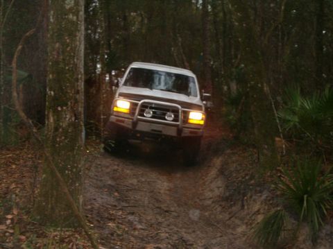 1992 Ford Bronco - A 1992 Ford Bronco navigates a wooded trail, its headlights illuminating the path ahead.