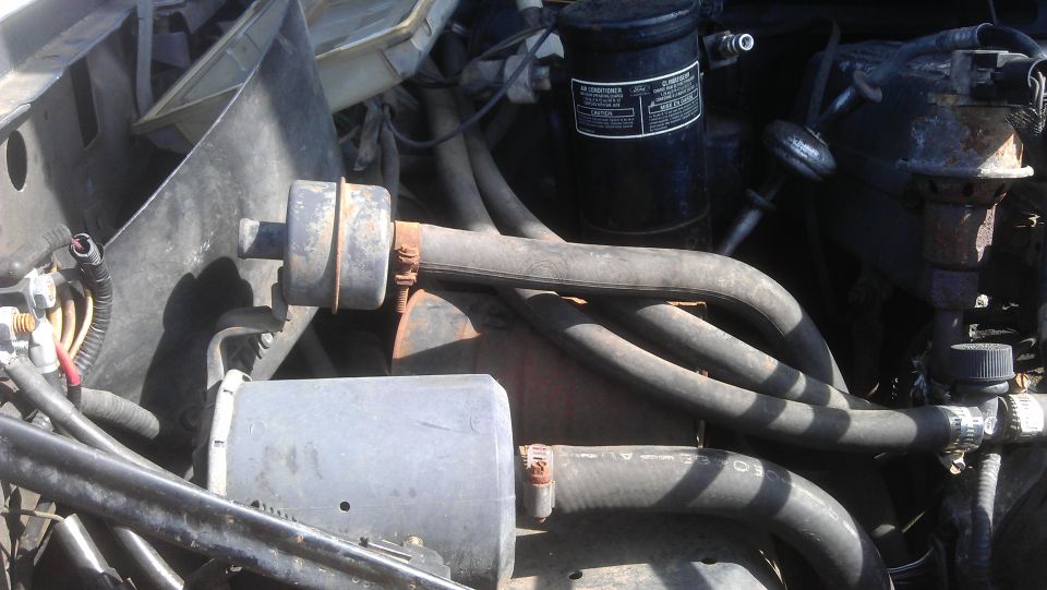 Engine bay with hoses, a black cylinder with a label, and metal components. Some rust is visible