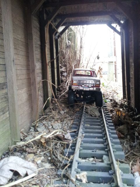 1995 Ford Bronco - A toy truck is positioned on a model train track within a rustic, overgrown tunnel.