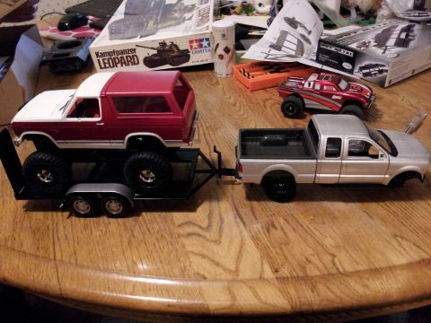 1995 Ford Bronco - A toy pickup truck tows a toy SUV on a trailer, surrounded by model kits on a wooden table.