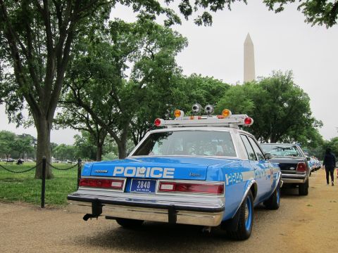 1991 Ford LTD Crown Victoria - A line of police cars, including a blue Dodge, are parked outdoors with trees and the Washington Monument in the background.