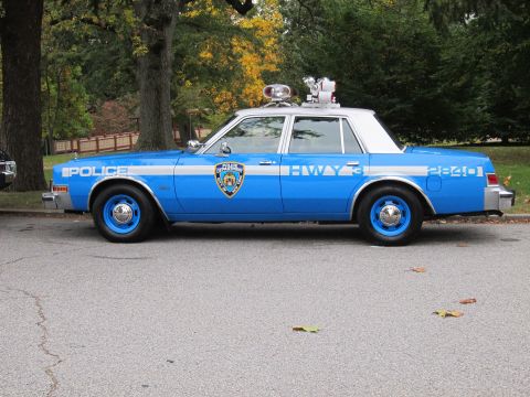 1991 Ford LTD Crown Victoria - Side profile of a blue-and-white NYPD-liveried Dodge Diplomat four-door sedan on pavement under autumn trees. It wears a white roof, silver…