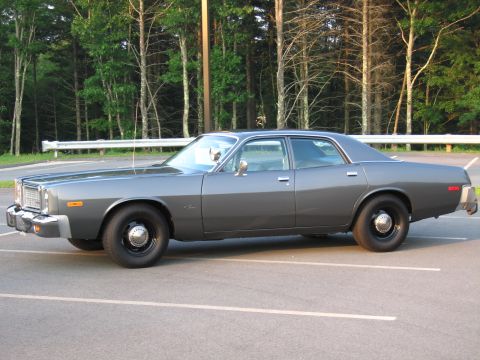 1991 Ford LTD Crown Victoria - Gray full-size Mopar four-door sedan photographed in profile in an empty parking lot backed by woods and guardrail. It has a plain unmarked…