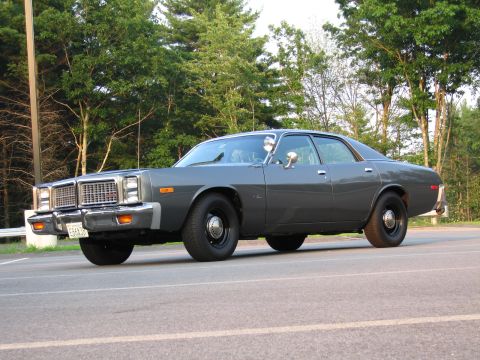 1991 Ford LTD Crown Victoria - Gray late-1970s Dodge Monaco four-door sedan parked at a 3/4 front angle in an empty lot bordered by guardrail and trees. It has a plain un…