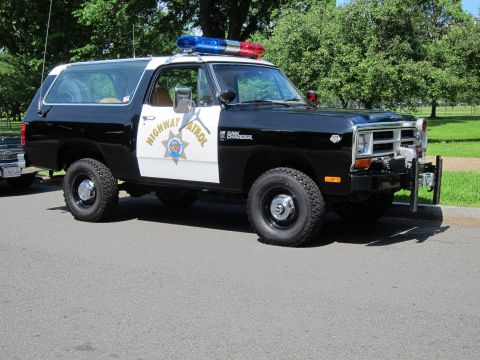1991 Ford LTD Crown Victoria - Black-and-white Dodge Ram Charger 4x4 in California Highway Patrol livery, shot from a 3/4 front angle on pavement beside a grassy park. It…