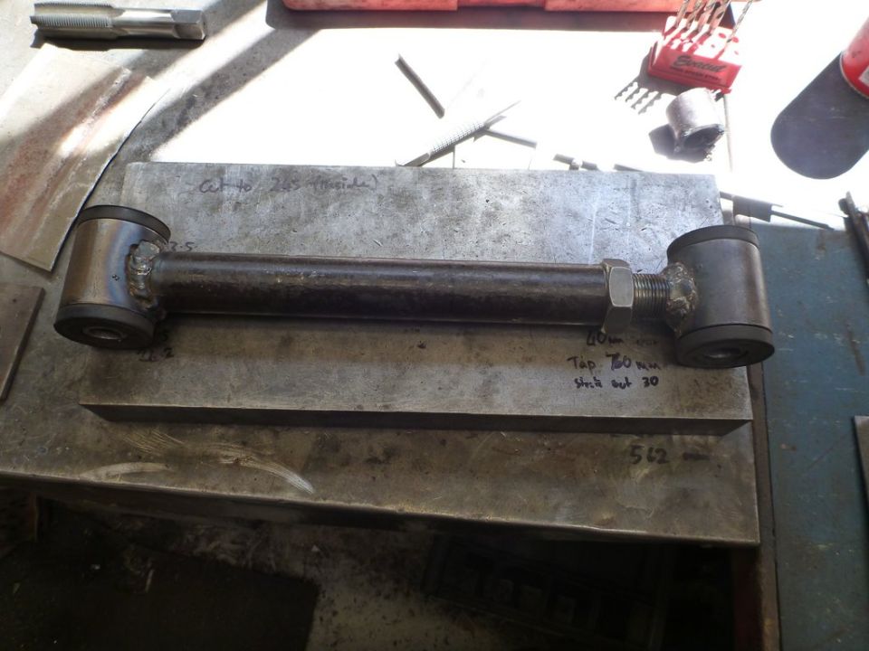 Custom suspension control arm with rubber bushings and a nut on a threaded end, resting on a metal surface with handwritten notes