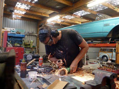 1964 Mercury Monterey - A man works on metal with an angle grinder in a workshop, while a vintage blue Mercury Monterey convertible is suspended above.