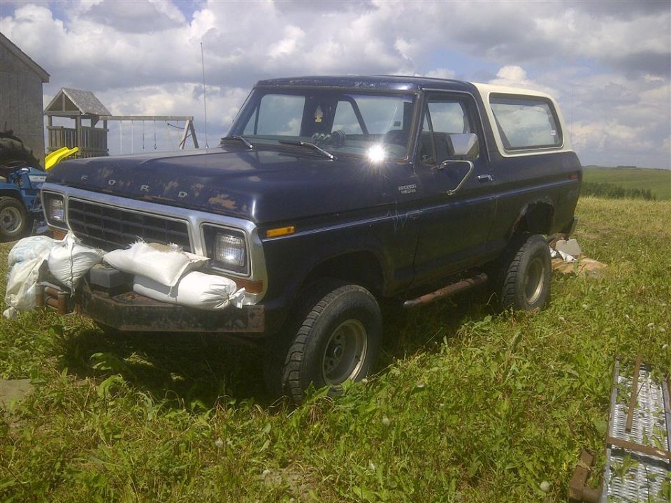 Dark blue Ford Bronco with a white top parked in tall grass. Bags of sand are on the front bumper