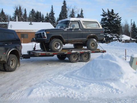 1979 Ford Bronco - A dark gray Ford Bronco is loaded onto a trailer, being towed by a black truck through a snowy landscape.