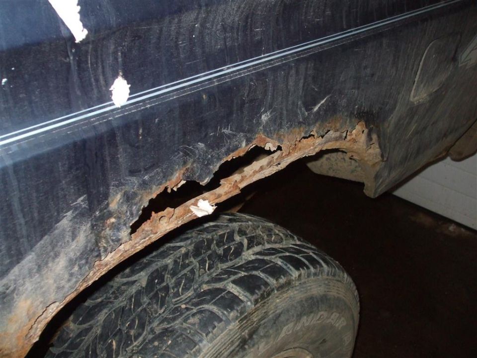 Close-up of severe rust damage on the rear wheel well of a dark-colored vehicle, with a tire visible below