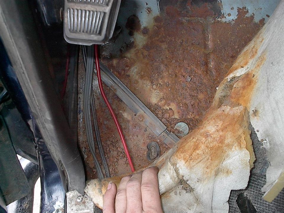 Close-up of a rusty floor pan inside a vehicle, with wiring and part of a pedal visible