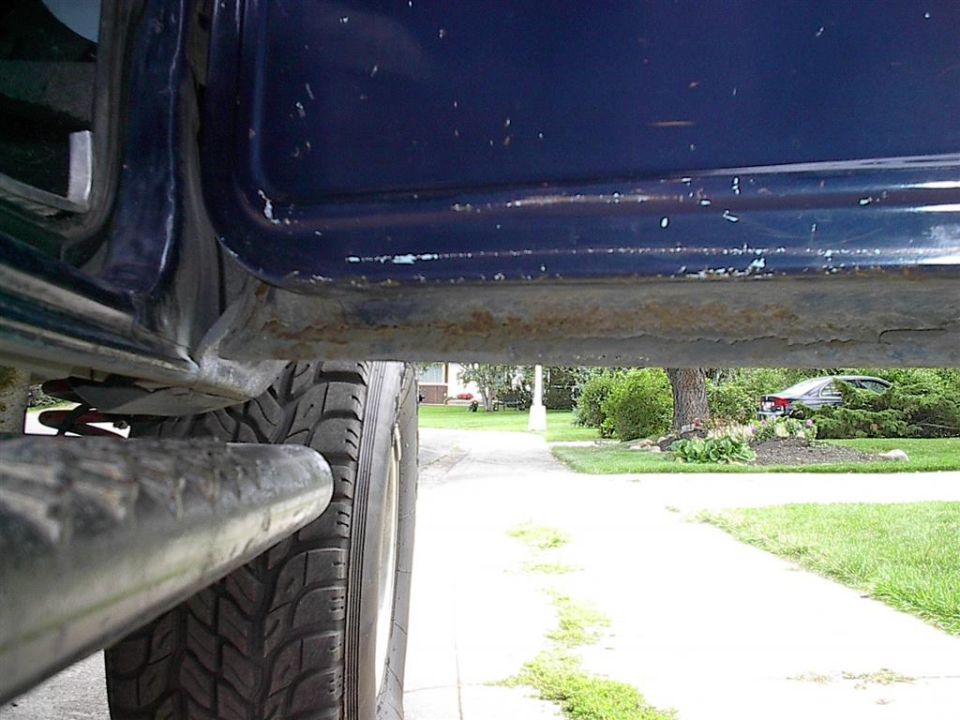 Underside view of a dark blue vehicle's rocker panel showing rust, with a large tire in the foreground. Another car is visible in the background