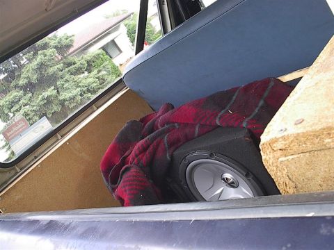 1979 Ford Bronco - A subwoofer and blanket are stored in the cargo area of a vehicle, with a view of trees and a house through the window.