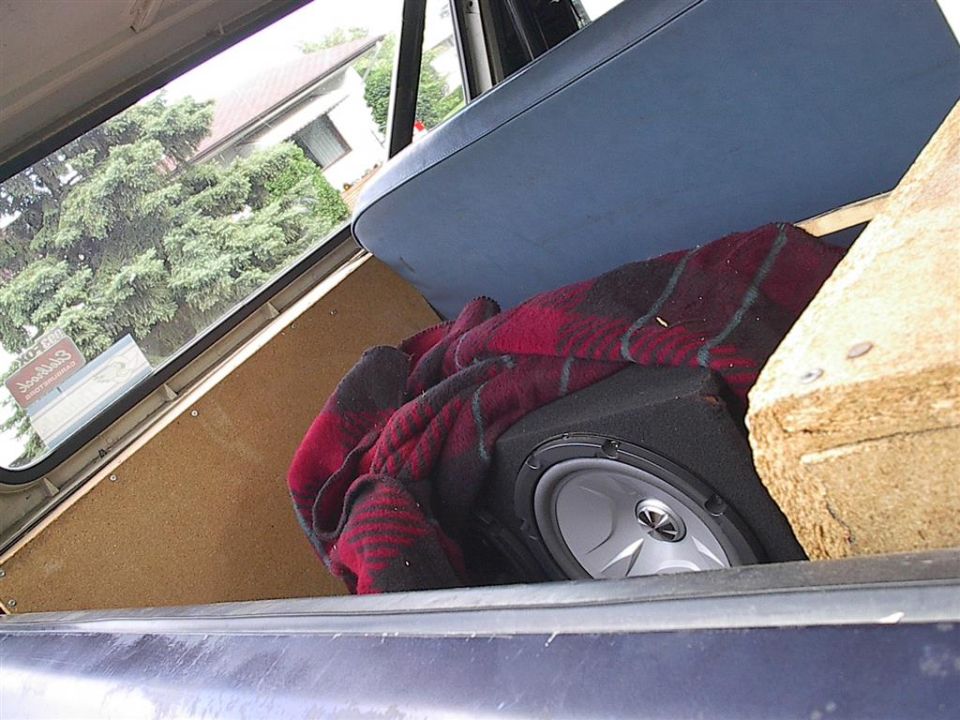 Inside a vehicle, looking towards the back. There's a subwoofer covered by a red and black plaid blanket, and a blue seat is visible