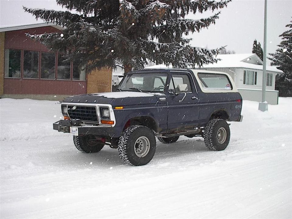 Dark blue 1978 Ford Bronco with a white removable top, lifted suspension, and off-road tires parked in snow