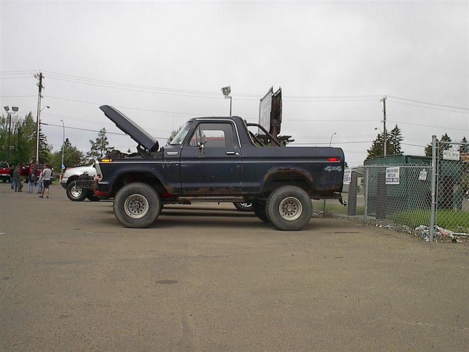 Dark blue Ford Bronco with hood up, parked next to a white truck. 4x4 decal on rear fender