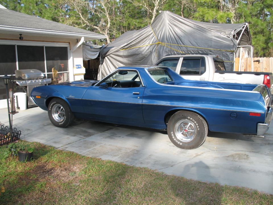 Blue 1972 Ford Ranchero parked in driveway next to a white pickup truck under a tarp